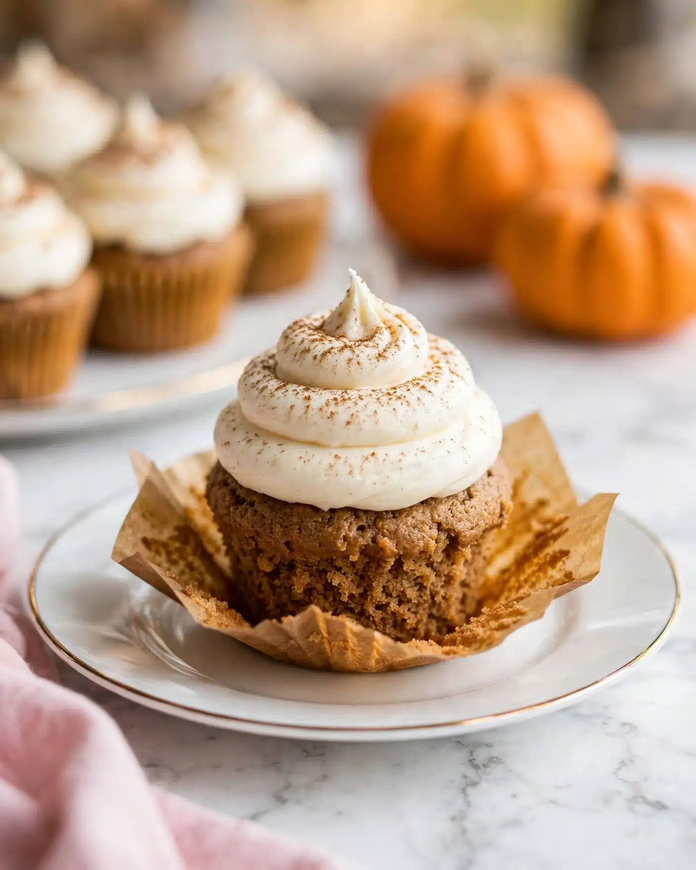 Pumpkin Cupcakes with Cream Cheese Frosting Recipe 5 A single cupcake sits in the center on a white plate with a thin gold rim, placed on a white marbled surface. The cupcake has two layers: the bottom is a dense, light brown cake with a slightly rough texture, partially wrapped in an unwrapped paper liner that is golden brown and flares out around the base. The top layer is a tall, smooth swirl of creamy white frosting, piped in a spiral peak that sits neatly on the cake. The frosting is lightly dusted with a fine brown powder, adding a subtle speckled texture. In the background, out of focus, are more similar cupcakes on a white plate and two small pumpkins in warm orange tones, creating a cozy autumn feel. The edges of a pink cloth can also be seen at the bottom left corner of the image, adding softness to the composition. photo taken with an iphone --ar 4:5 --v 7