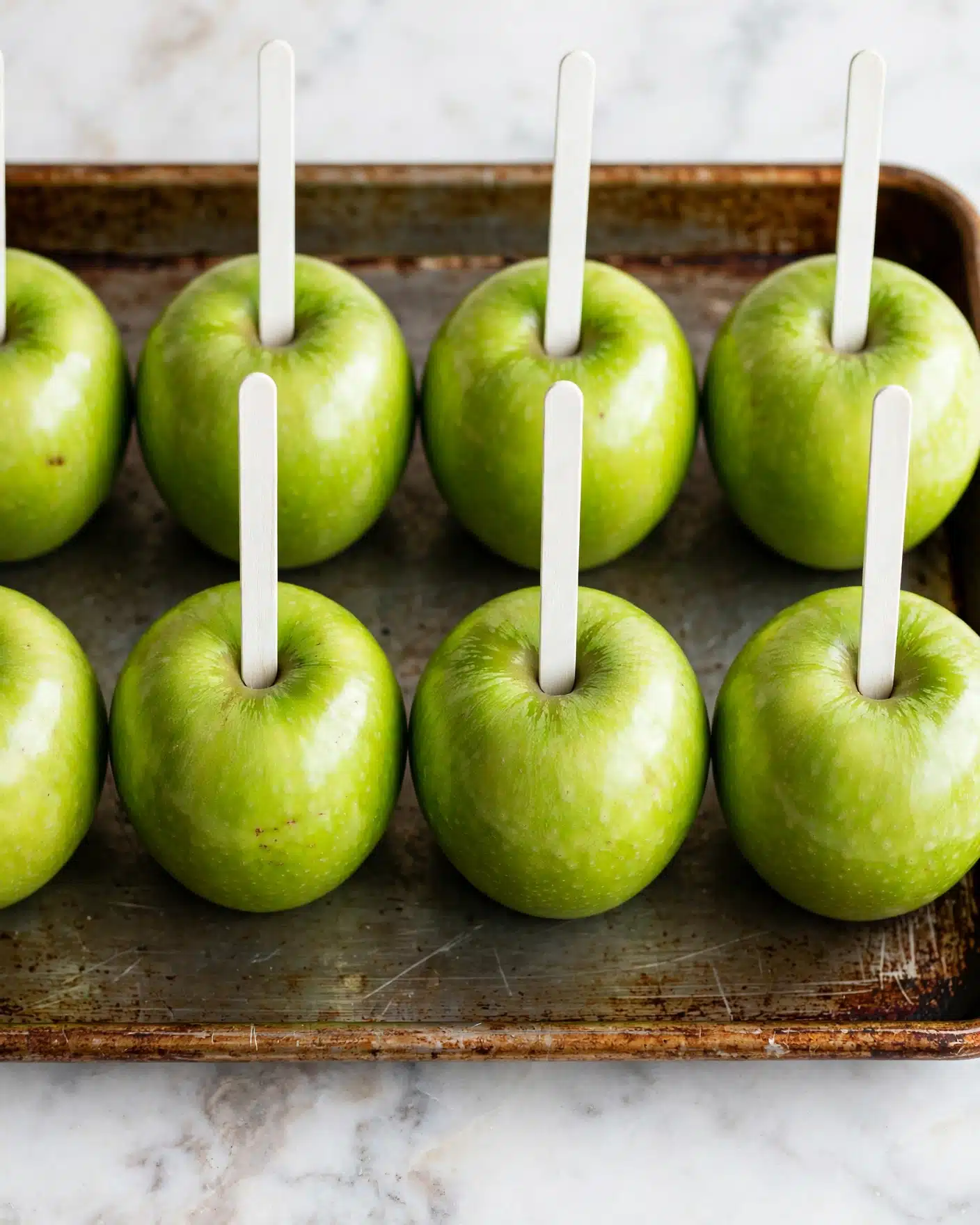 Homemade Caramel Apples Recipe 4 A close-up view of two rows of bright green apples, each with a white stick inserted straight into the top center. The apples sit evenly spaced on a well-used metal tray with a slightly rusty, brownish edge. The background and surface beneath the tray have a smooth white marbled texture, making the green apples stand out in sharp contrast. The lighting is soft and natural, highlighting the shiny, smooth skins of the apples, with the sticks rising vertically from them. photo taken with an iphone --ar 4:5 --v 7
