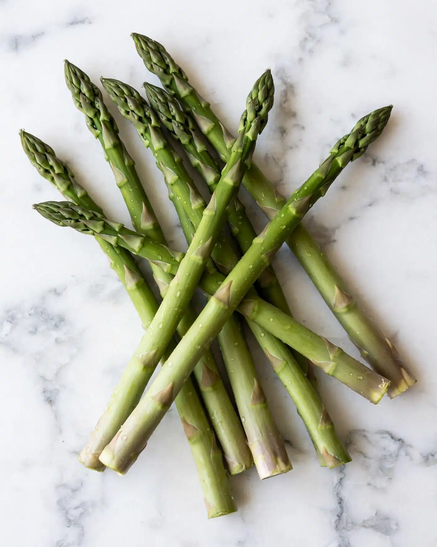 One-Pan Garlic Butter Chicken with Asparagus Recipe 4 A pile of fresh green asparagus spears lay on a white marbled surface, with each spear showing a smooth, slightly glossy texture. The asparagus stalks are firm and long, arranged loosely in a small stack with the tips pointing in different directions. The tips are tightly closed and have a slightly darker green color compared to the lighter green of the stalks. The overall look is clean and fresh with no additional items in the scene. photo taken with an iphone --ar 4:5 --v 7