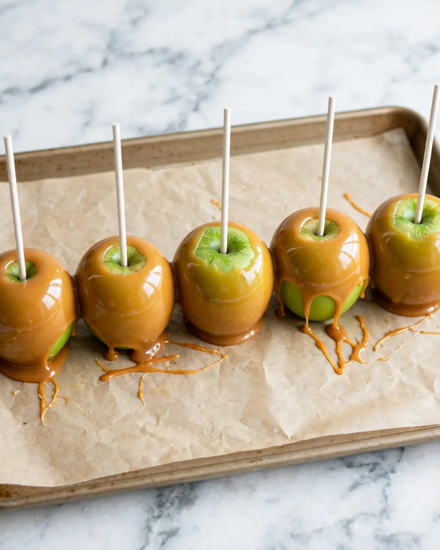 Homemade Caramel Apples Recipe 5 A row of shiny green apples each covered in a smooth, thick caramel layer with a glossy finish, placed on a light brown baking sheet lined with parchment paper. Each apple has a white stick inserted straight into the center top, sticking up tall. The apples show a clear green patch near the top where the caramel does not fully cover. The background is a white marbled surface that softly contrasts with the warm caramel color. Photo taken with an iphone --ar 4:5 --v 7