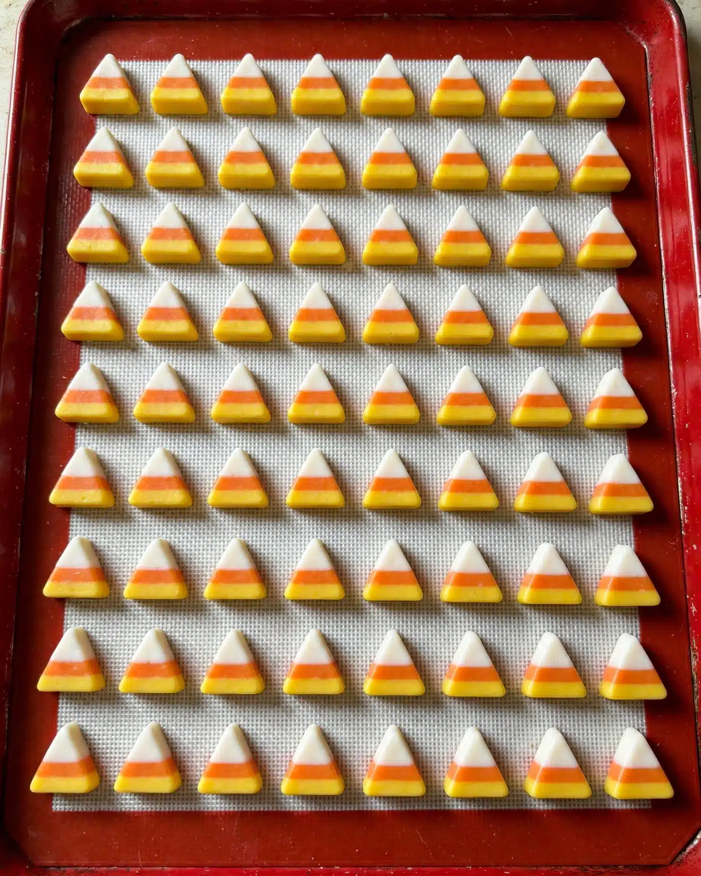Candy Corn Cookies Recipe 4 The image shows many small candy pieces shaped like triangles with three clear layers, arranged neatly in rows on a silicone mat inside a red baking tray. Each candy piece has a bright yellow bottom layer, a middle orange layer, and a top white layer, with smooth textures. The candies are evenly spaced with slight shadows visible on the white silicone mat, creating a tidy and colorful pattern. The photo is taken from above with soft lighting highlighting the candy layers and the red tray edges. photo taken with an iphone --ar 4:5 --v 7