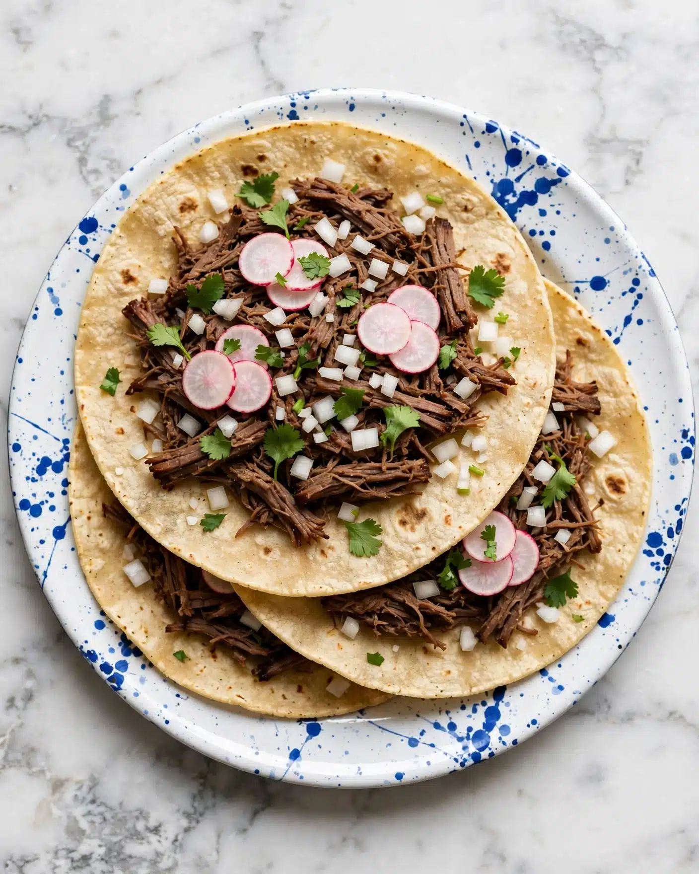 Slow Cooker Barbacoa Beef Recipe 5 Three soft corn tortillas stacked slightly overlapping on a white plate with blue splatter patterns, each filled with shredded dark brown meat in the center as the thickest layer. On top of the meat are thin, round slices of pale pink radishes arranged in small groups, along with small white diced onions scattered evenly. Bright green cilantro leaves sprinkled throughout add a fresh touch. The white marbled surface under the plate contrasts with the vibrant colors of the tacos. photo taken with an iphone --ar 4:5 --v 7