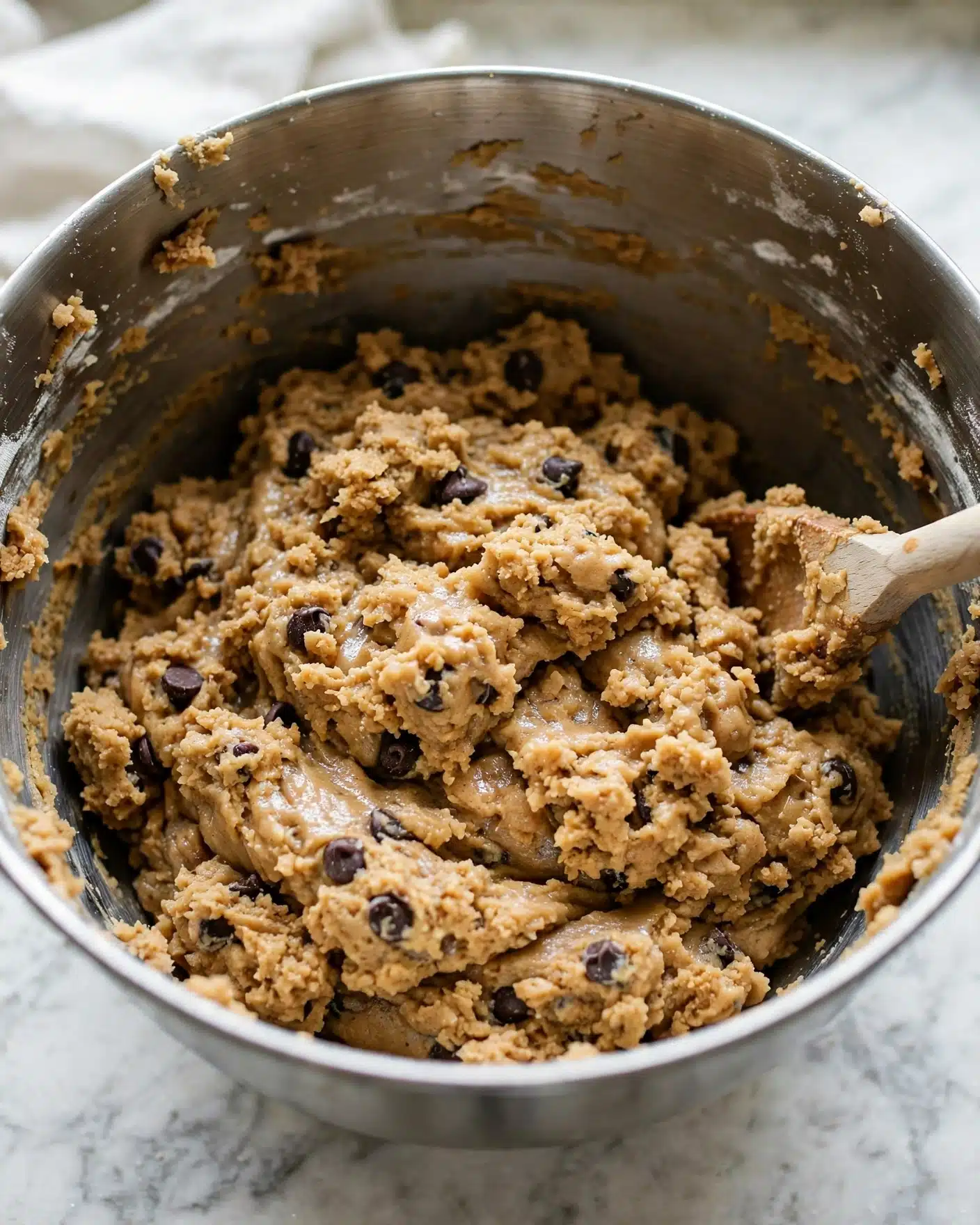 Jack-O-Lantern Pumpkin Cookies Recipe 4 A large metal mixing bowl filled with thick, tan cookie dough that has visible dark chocolate chips scattered throughout. The dough has a slightly rough but moist texture, piled mostly in the center and sticking somewhat to the sides of the bowl. A wooden spoon with a pale handle is partially inserted in the dough on the right side, and bits of dough are stuck to the inner edges of the bowl. The bowl sits on a white marbled surface, and natural light adds a soft shine to the dough. photo taken with an iphone --ar 4:5 --v 7