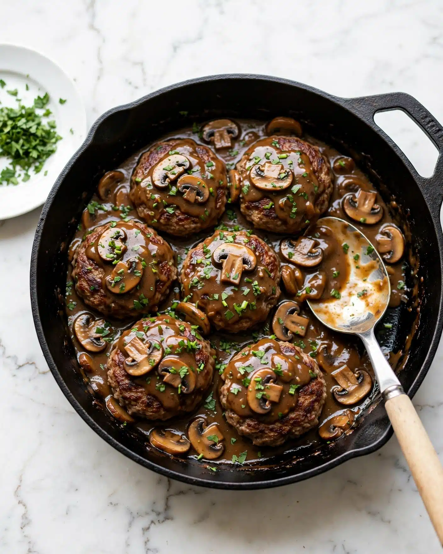 Classic Salisbury Steak with Mushroom Gravy Recipe 5 The image shows a black cast iron pan filled with six browned, round meat patties covered in a thick brown mushroom sauce. The sauce includes sliced mushrooms and small green herb pieces sprinkled over the top, adding texture and color. A silver spoon with a beige wooden handle rests in the pan’s right side, partially covered in the sauce. To the left of the pan, there is a small light wood plate with finely chopped green herbs on a white marbled surface. photo taken with an iphone --ar 4:5 --v 7