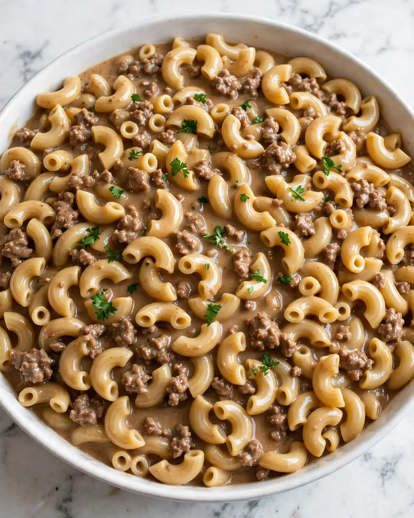 Creamy Hamburger Helper Recipe 5 A close-up view of creamy elbow macaroni pasta mixed evenly with browned ground meat, all coated in a smooth, light brown sauce with a slightly glossy texture. Small green parsley bits are sprinkled on top, adding a fresh color contrast to the warm tones of the dish. The food fills a large white bowl that sits on a white marbled surface. The overall look is rich and comforting, showing the soft, curved pasta shapes nestled tightly with tender meat pieces in the sauce. Photo taken with an iphone --ar 4:5 --v 7