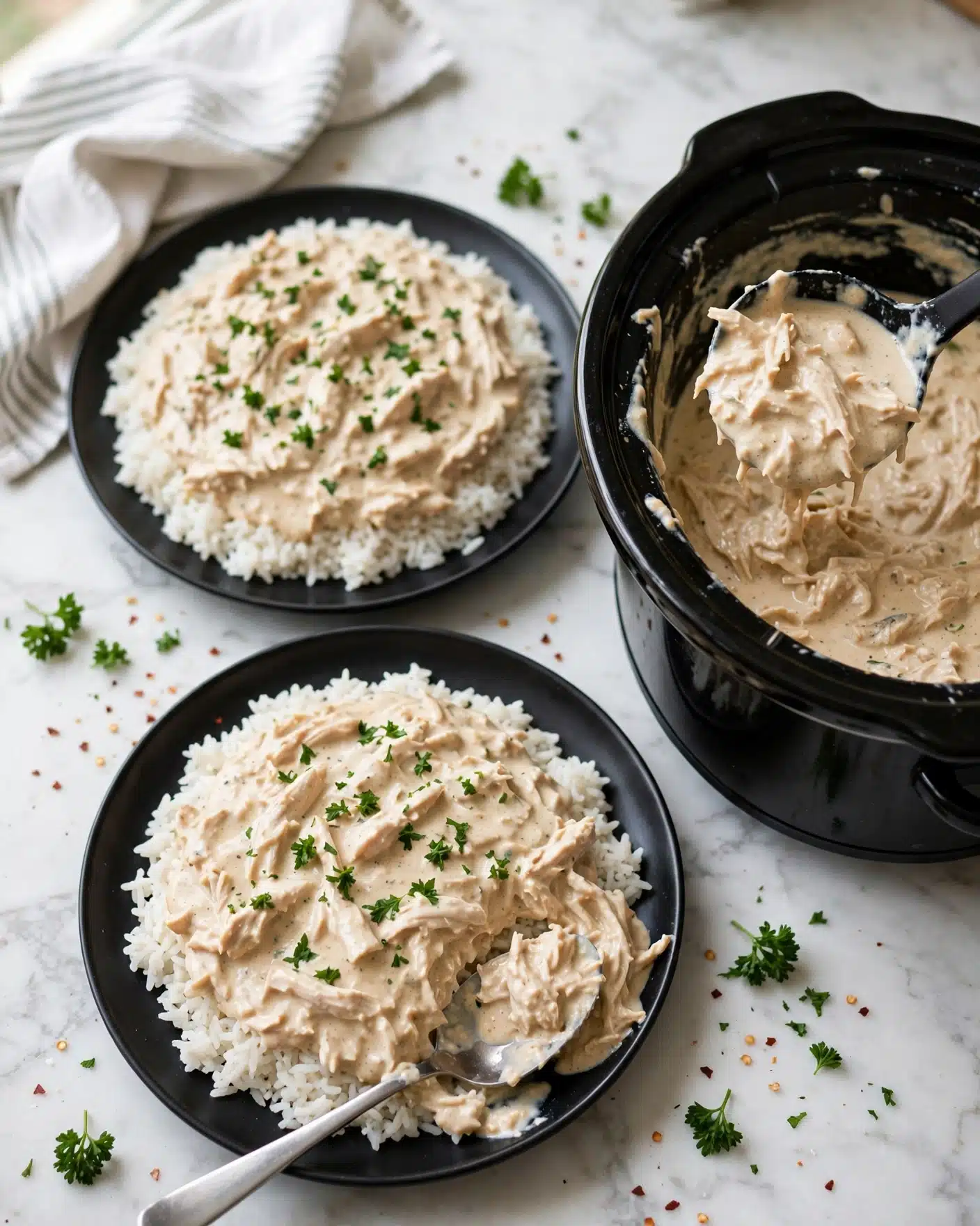 Crockpot Chicken and Gravy Recipe 5 Two black plates on a white marbled surface each hold a layer of white rice spread wide, topped with a creamy light beige sauce mixed with shredded chicken. Small green parsley pieces sprinkle over the sauce, adding bright green color. A silver spoon rests on the lower plate partly digging into the sauce. To the right, a black slow cooker filled with more creamy chicken and sauce sits open, with a ladle lifting some of the sauce. Some parsley sprigs and light pepper flakes are scattered around the plates. A white cloth with stripes lies near the top left corner. Photo taken with an iphone --ar 4:5 --v 7