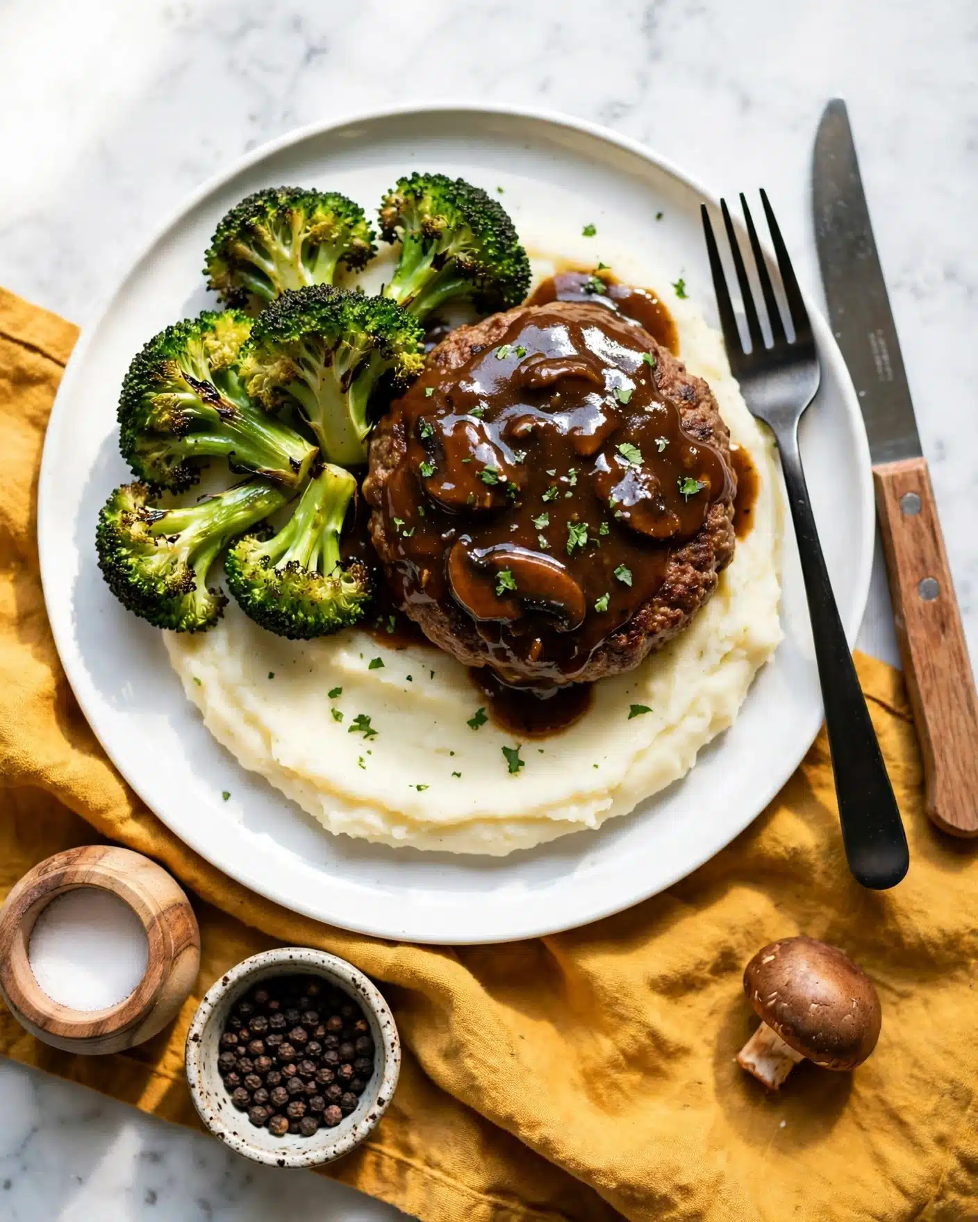 Easy Homemade Salisbury Steak Recipe 5 A white plate with three main layers: at the bottom is a smooth, creamy white layer of mashed potatoes; on top of it sits a thick, brown patty covered with a glossy, dark brown mushroom sauce sprinkled with small green herb pieces; to the left of the patty are several bright green, roasted broccoli florets with slightly charred edges. A black fork and a knife with a wooden handle rest on the right side of the plate. The plate is placed on a mustard yellow cloth on a white marbled surface. Nearby, there is a small wooden salt container, a bowl with black pepper, and a single brown mushroom. photo taken with an iphone --ar 4:5 --v 7