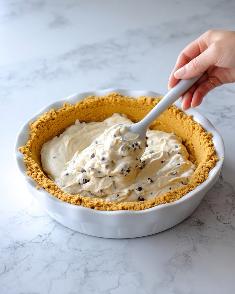 A white pie dish holds a thick, crumbly, golden-brown crust layer evenly pressed inside. A woman's hand is using a white spatula to scoop and spread a creamy, light beige filling mixed with small dark chocolate chips from a pale grey mixing bowl into the crust. The background is a white marbled surface with faint grey veining, enhancing the light and clean appearance of the scene. The filling has a smooth, fluffy texture with chocolate bits spread throughout. Photo taken with an iphone --ar 4:5 --v 7