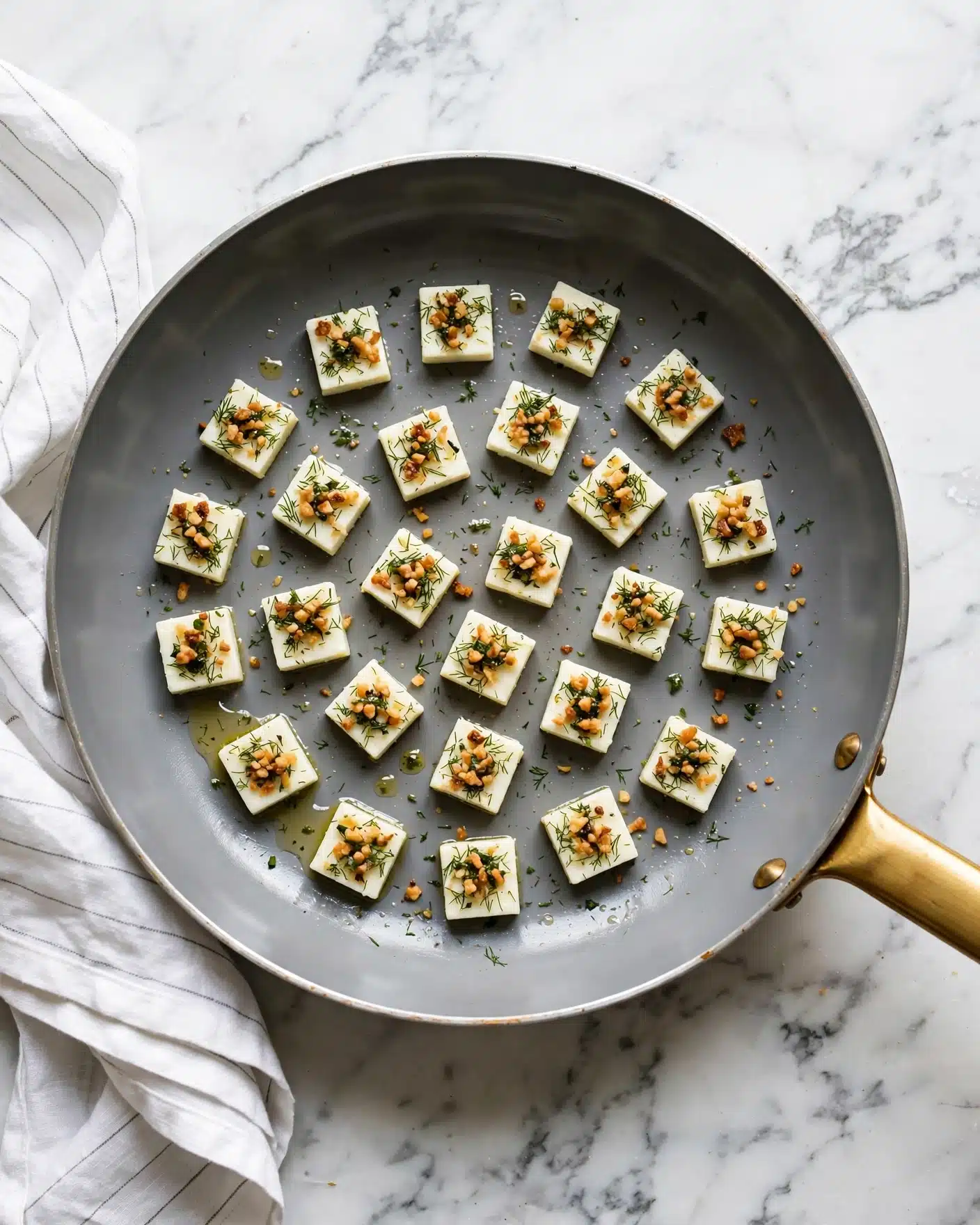 A gray frying pan with a golden handle holds about 24 small square pieces of white cheese, each piece topped with a mix of greenish herbs and golden brown bits that look crispy. The cheese pieces are arranged loosely in the pan with some space between them, and a few drops of oil and small herb crumbs are scattered around. The pan rests on a white marbled surface with a white cloth that has thin gray lines folded to the left side. The photo taken with an iphone --ar 4:5 --v 7