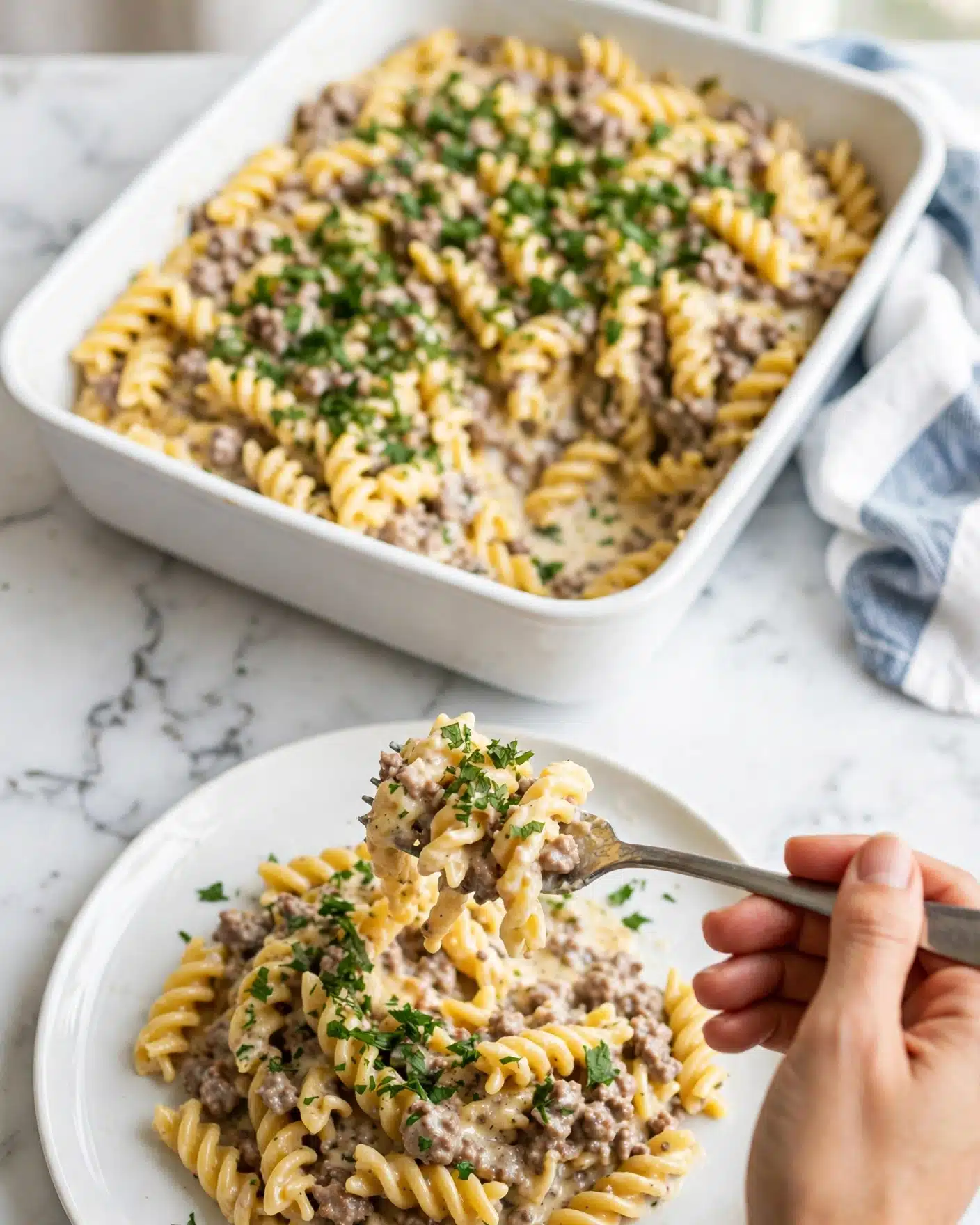A white baking dish filled with creamy pasta mixed with ground meat and topped with chopped green herbs is placed on a white marbled surface. In front of it, a white plate holds a serving of the same creamy pasta with meat, showing swirling yellowish pasta pieces mixed with light sauce and specks of meat, garnished with fresh green herbs. A woman's hand is holding a fork, lifting some pasta on the plate. The overall scene has a bright, clean look with a soft blue and white cloth partially visible near the baking dish. photo taken with an iphone --ar 4:5 --v 7