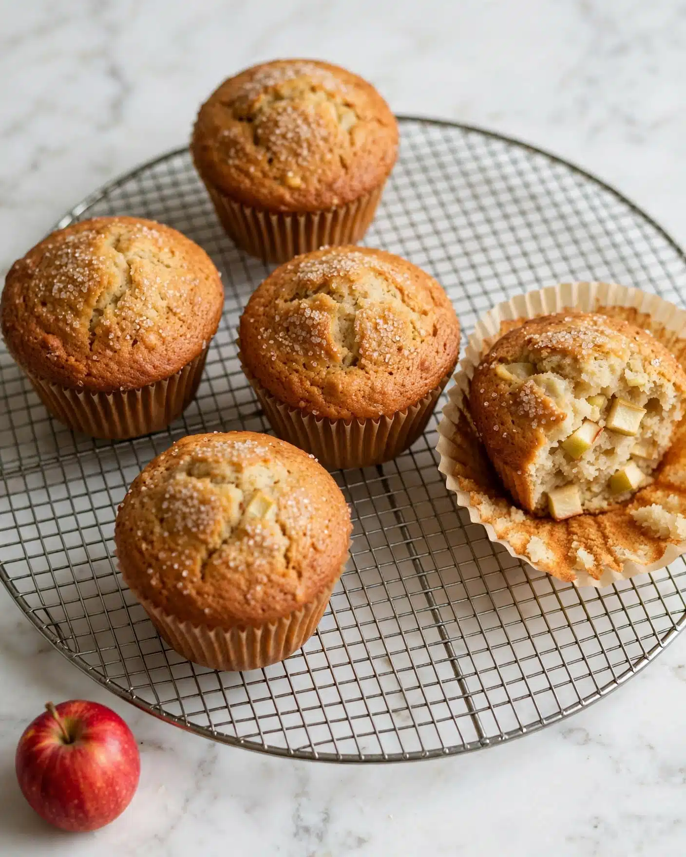 Gluten-Free Spiced Apple Muffins (Almond & Oat Flour) Recipe 5 The image shows five golden brown muffins on a round metal cooling rack over a white marbled surface. Each muffin has a slightly domed, textured top with a light dusting of sugar crystals, showing a crumbly and soft texture. The muffins are wrapped in classic brown paper liners. One muffin on the right is partially unwrapped, revealing a pale moist inside with small chunks of apple visible. In the front left corner, there is a small red apple adding color contrast. The overall scene is warm and inviting, focused closely on the baked muffins. photo taken with an iphone --ar 4:5 --v 7