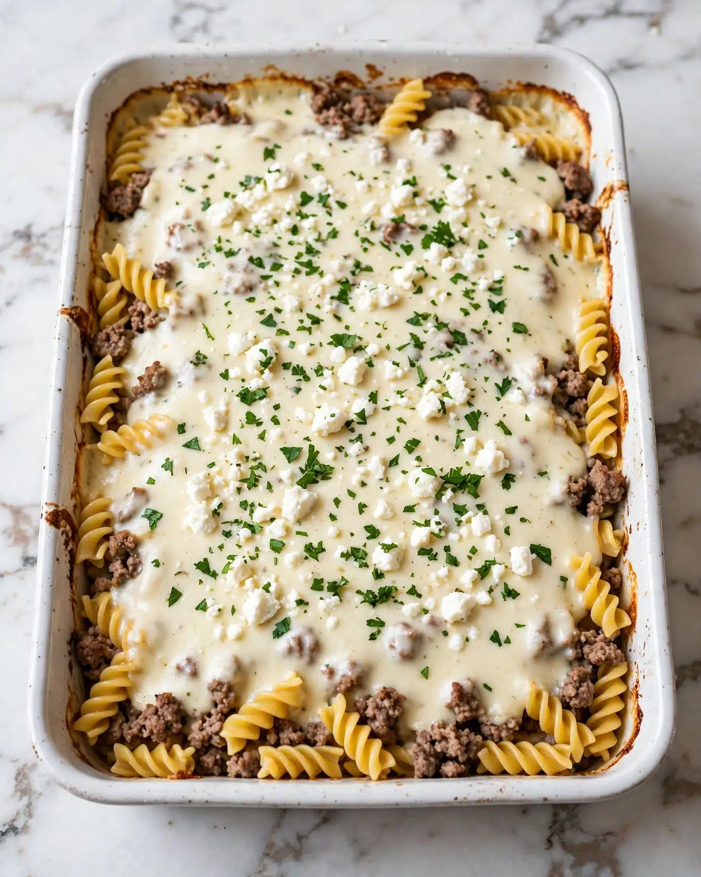 The image shows a rectangular white baking dish filled with a layered casserole. The base layer consists of twisted golden-yellow pasta intertwined with browned pieces of ground meat. On top, there is a creamy light-colored sauce evenly covering the pasta and meat. Sprinkled over the top are small white cheese crumbles and chopped green herbs, adding a fresh touch of color. The edges of the dish show some slight browning from baking. The dish is placed on a white marbled surface. Photo taken with an iphone --ar 4:5 --v 7