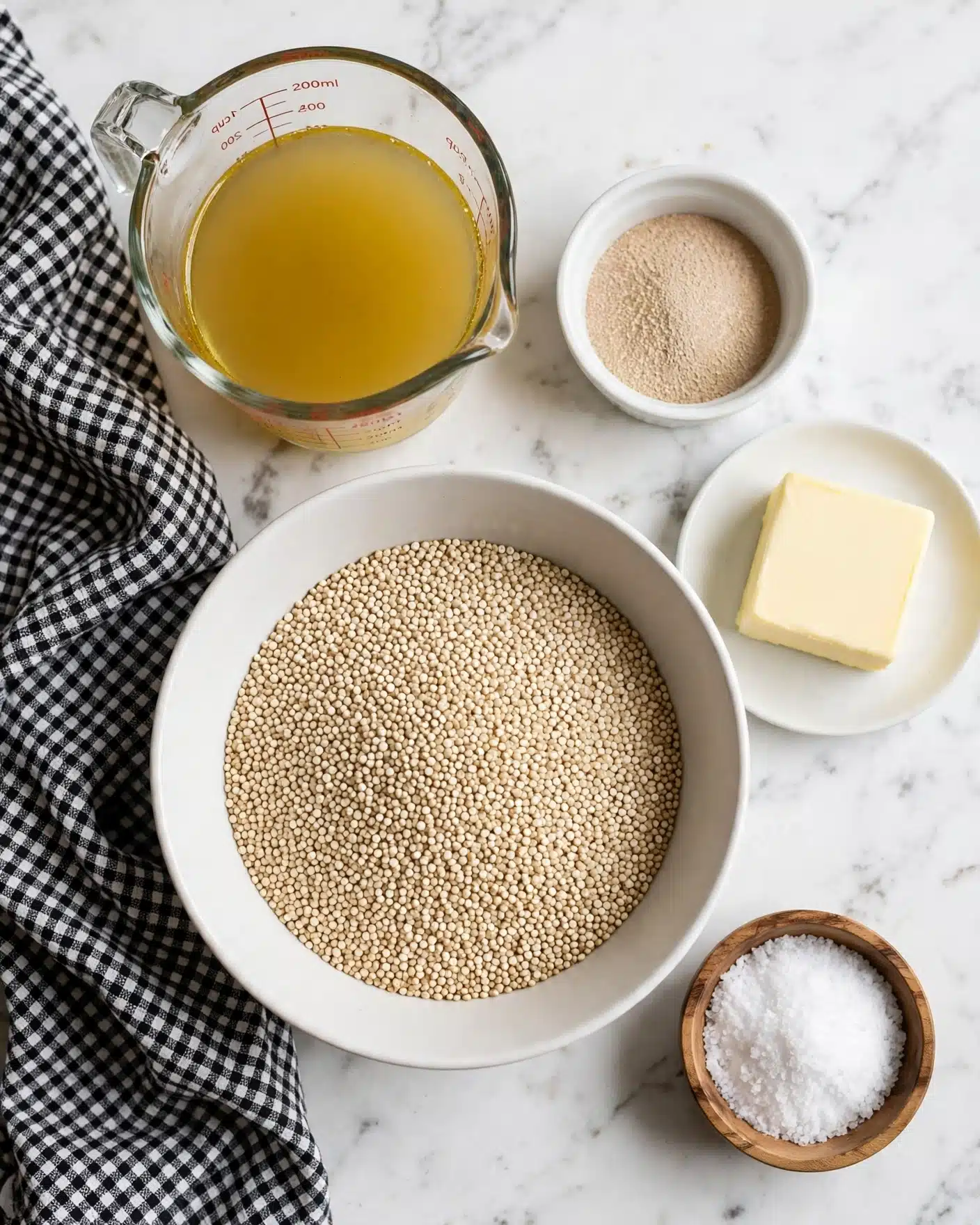 5-Ingredient Garlic Butter Quinoa Recipe 4 This image shows five different items on a white marbled surface. In the center, there is a large white bowl filled with small, round beige quinoa grains. Above it to the left, a clear glass measuring cup holds a yellow liquid, likely broth. To the right of the measuring cup, a small white bowl contains light brown granulated yeast. Below the yeast, a white shallow plate holds a thick square piece of pale yellow butter. At the bottom right, a small wooden bowl holds white coarse salt. A black and white checkered cloth is draped on the left side of the image. Photo taken with an iphone --ar 4:5 --v 7