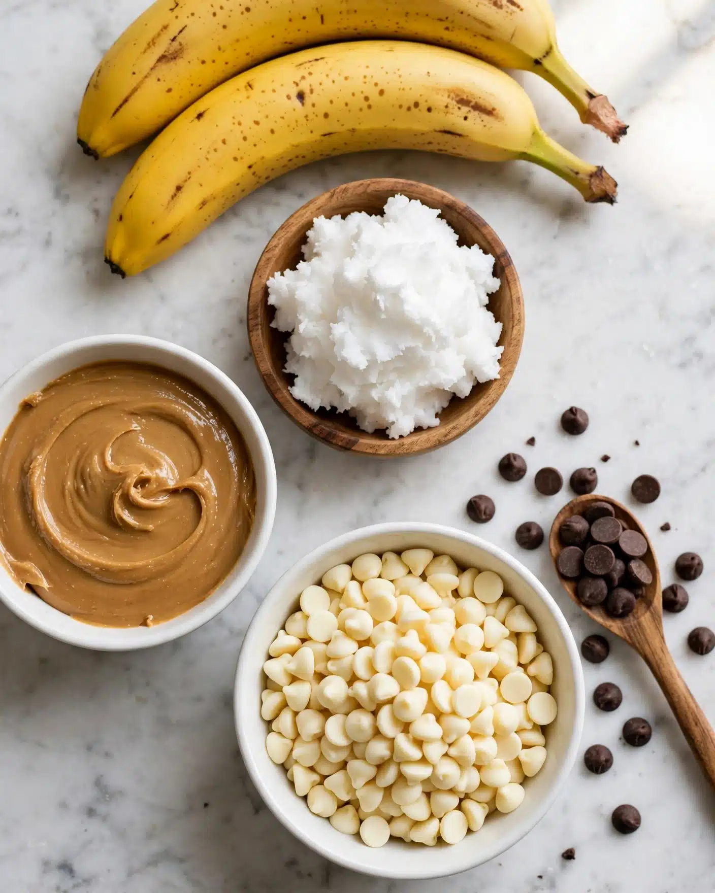 Chocolate Peanut Butter Banana Ghost Pops Recipe 4 The image shows ingredients arranged on a white marbled surface. At the top, there are two yellow bananas with small brown spots. Below the bananas, a small wooden bowl holds a dollop of white coconut oil with a soft, fluffy texture. To the left, there is a white bowl filled with smooth, light brown peanut butter. To the bottom right of the peanut butter, a larger white bowl is filled with creamy white chocolate chips, and next to it, a small wooden spoon holds dark brown chocolate chips with some scattered around. The photo taken with an iphone --ar 4:5 --v 7